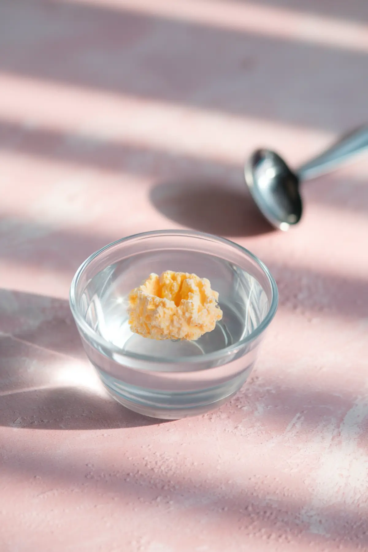Single piece of bloomed gelatin floating in a small clear bowl of water with soft morning light and shadows on a pink surface.
