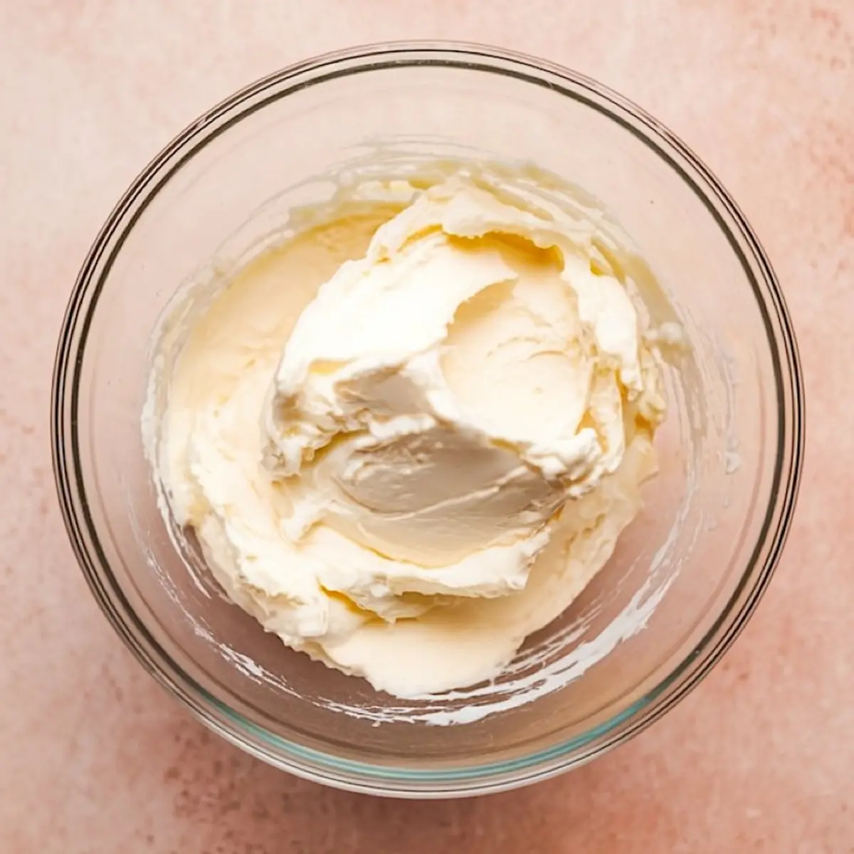 Overhead shot of a clear glass bowl with thick, creamy whipped topping or cream cheese mixture on a soft pink background.