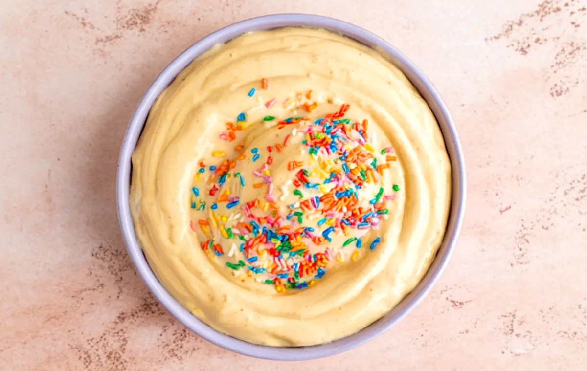 Bowl of creamy cake batter dip topped with colorful rainbow sprinkles on a light textured background.
