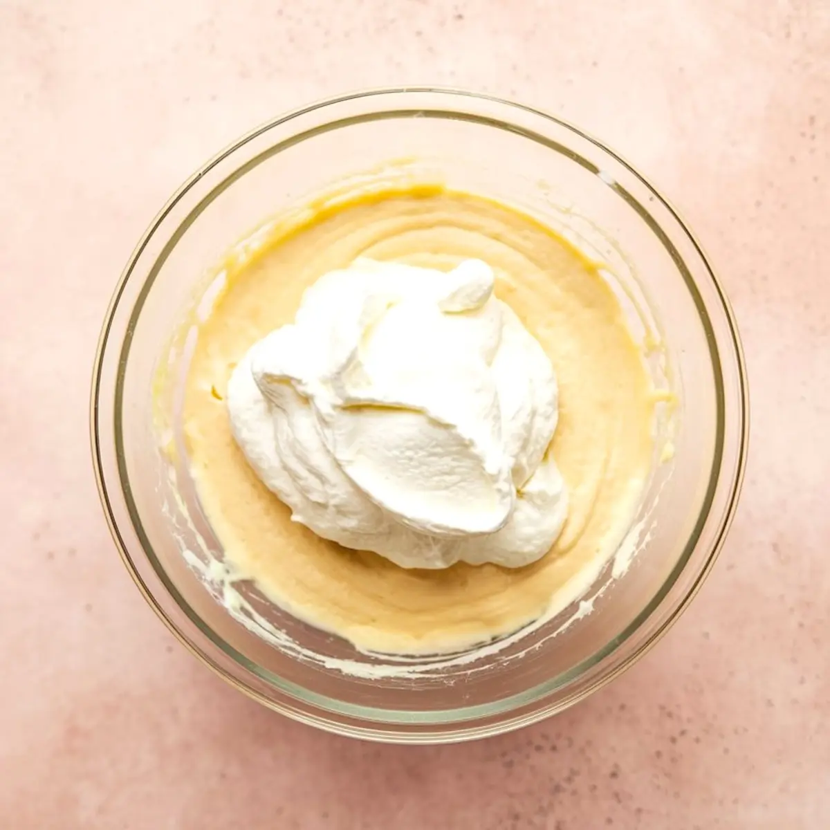 Glass bowl containing partially mixed cake batter with a large scoop of whipped topping on top, placed on a light pink surface.
