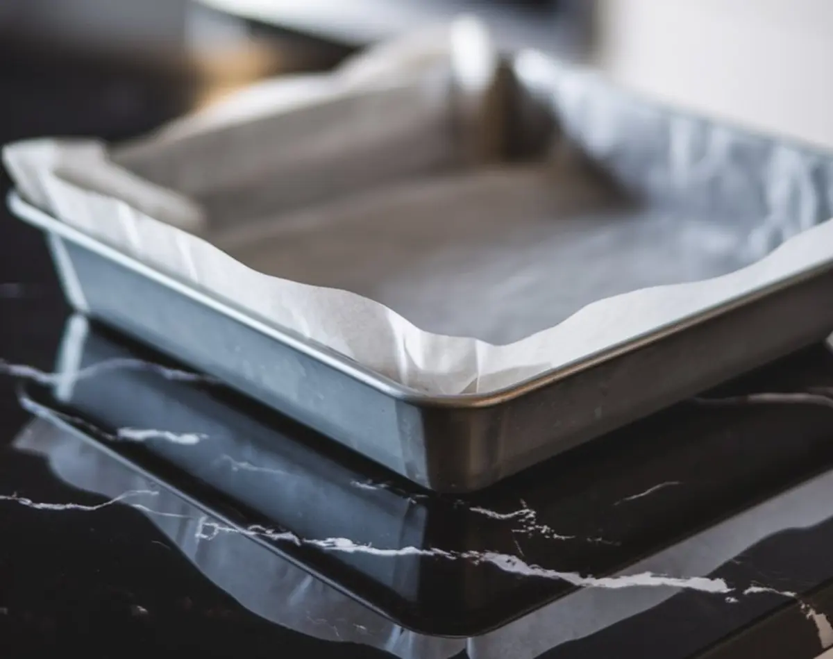 A square metal baking pan lined with white parchment paper, placed on a glossy black marble surface, ready for baking preparation.