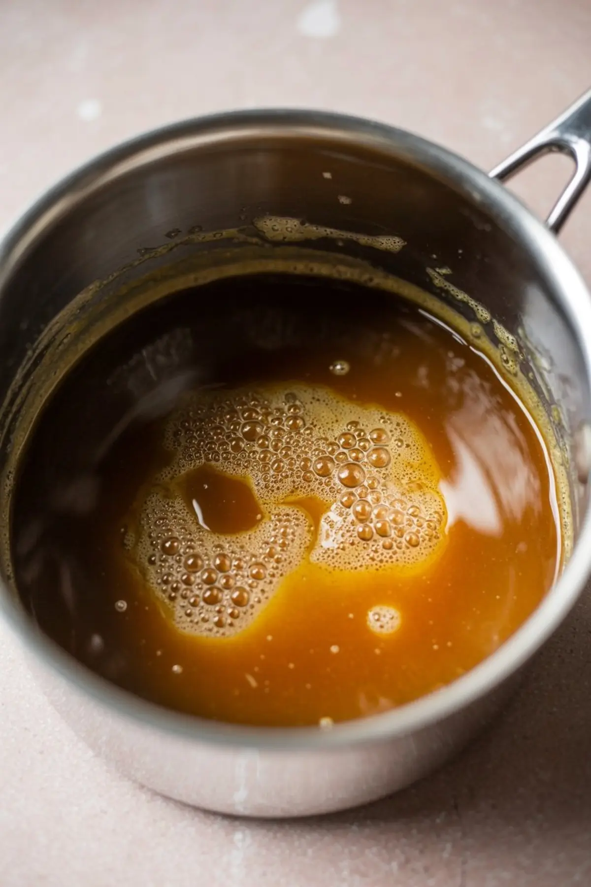 Golden caramel sauce bubbling in a metal saucepan on a light countertop, showing the mixture in early stages of cooking.
