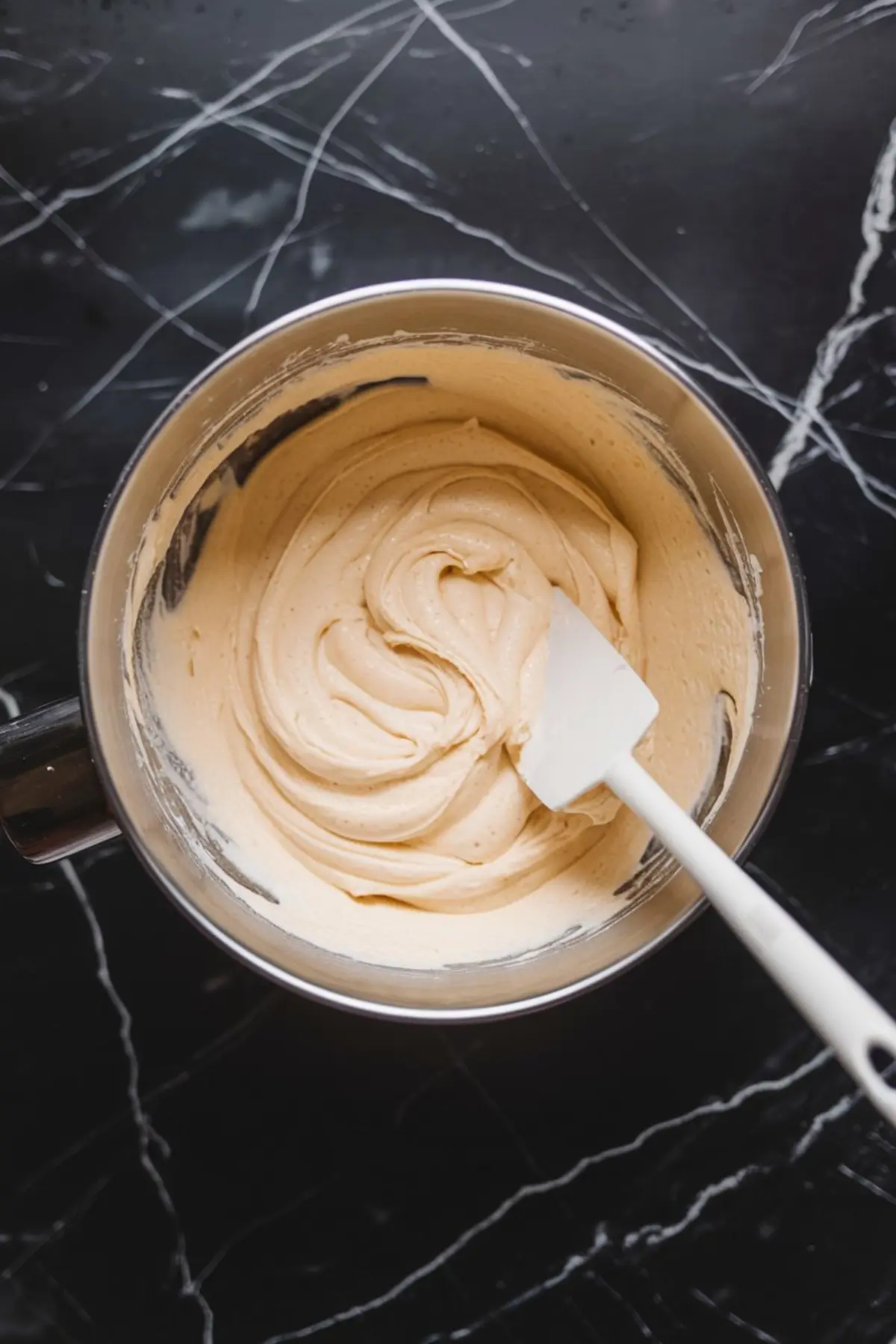 Mixing bowl filled with smooth cheesecake batter being stirred with a white spatula, on a black marble countertop.