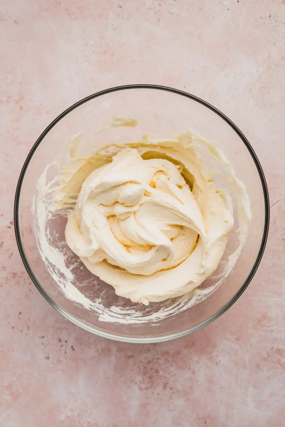 Whipped cream cheese mixture in a glass bowl on a light pink background, showcasing a smooth and fluffy texture ideal for dessert dips or frosting.