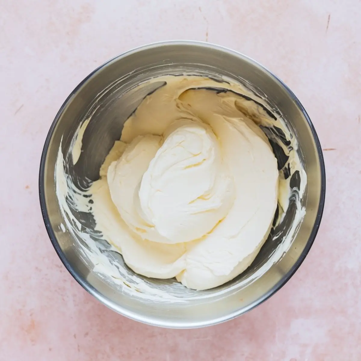 Fully mixed cheesecake filling with a silky, smooth texture inside a stainless steel mixing bowl, sitting on a light pink countertop.
