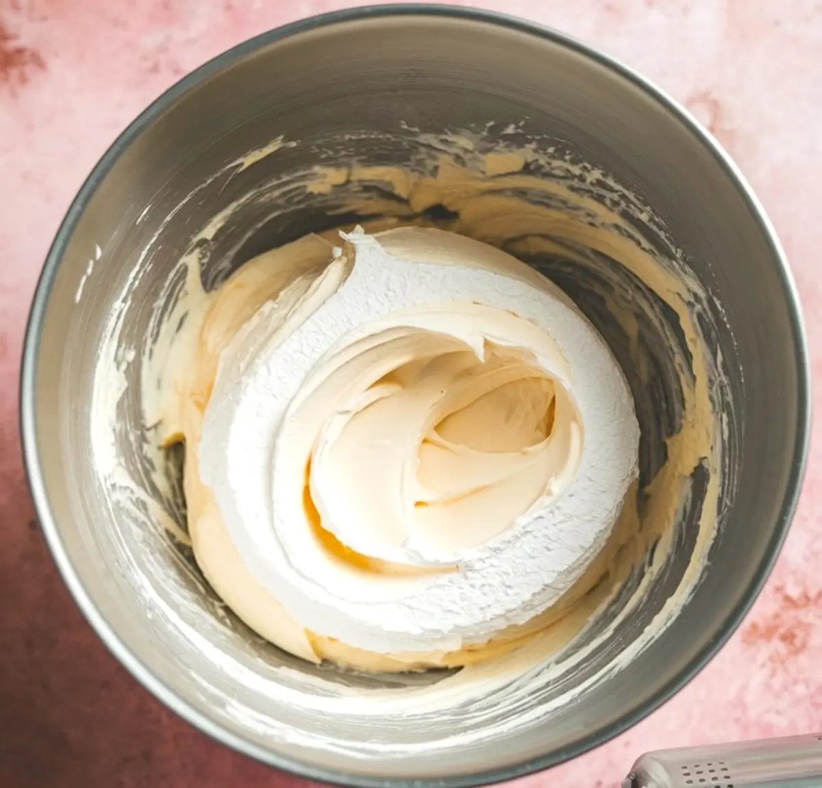 Cream cheese mixture swirled with powdered sugar in a metal mixing bowl, creating a smooth, creamy base for cheesecake dip, placed on a soft pink surface.

