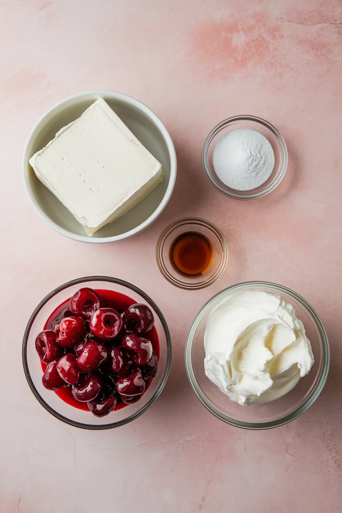 Flat lay of cheesecake dip ingredients on a pink background, including a block of cream cheese, granulated sugar, vanilla extract, whipped topping, and bright red cherries in syrup.
