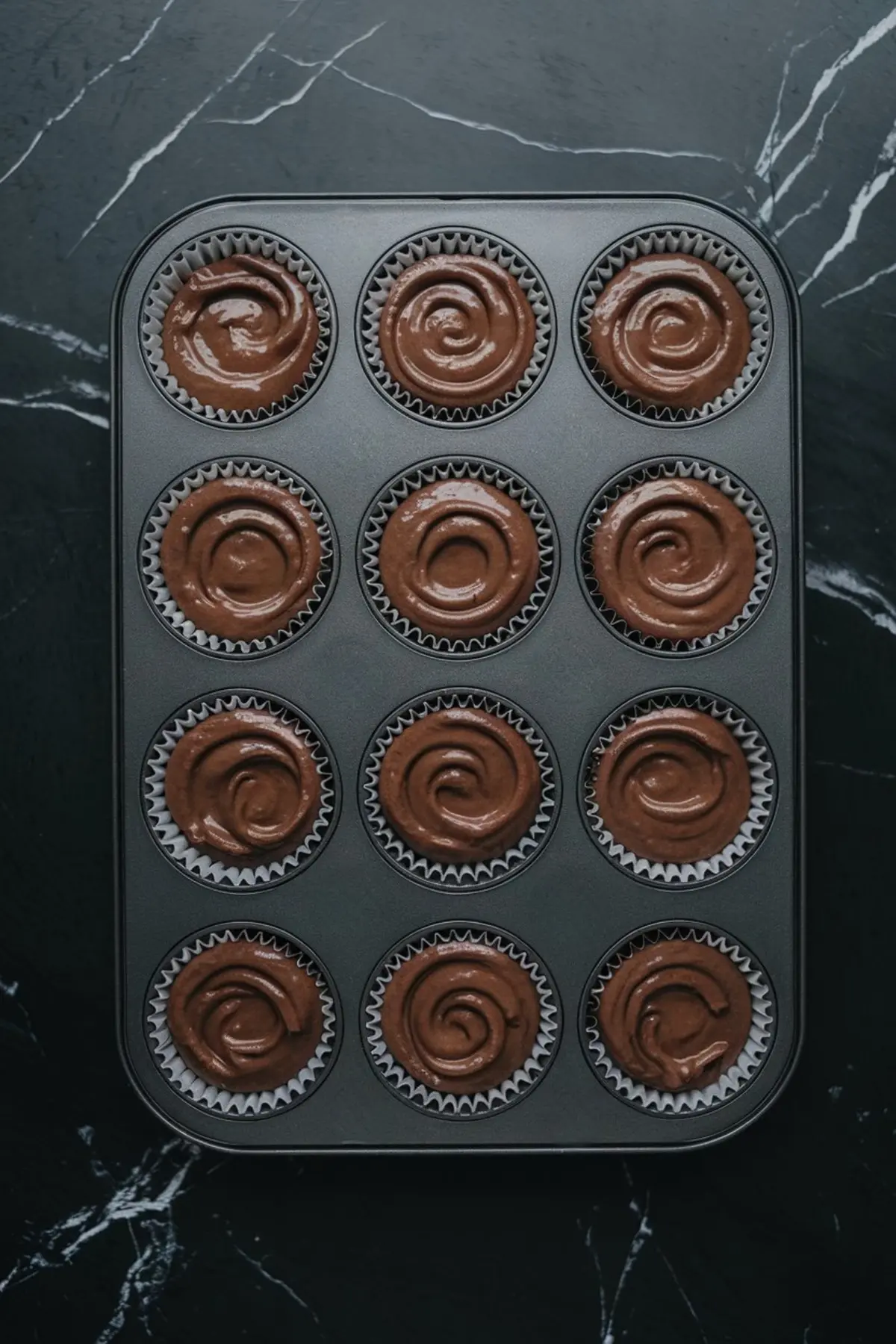 Top view of a 12-cup muffin pan filled with chocolate cupcake batter swirled in liners, ready for baking on a black marble surface.