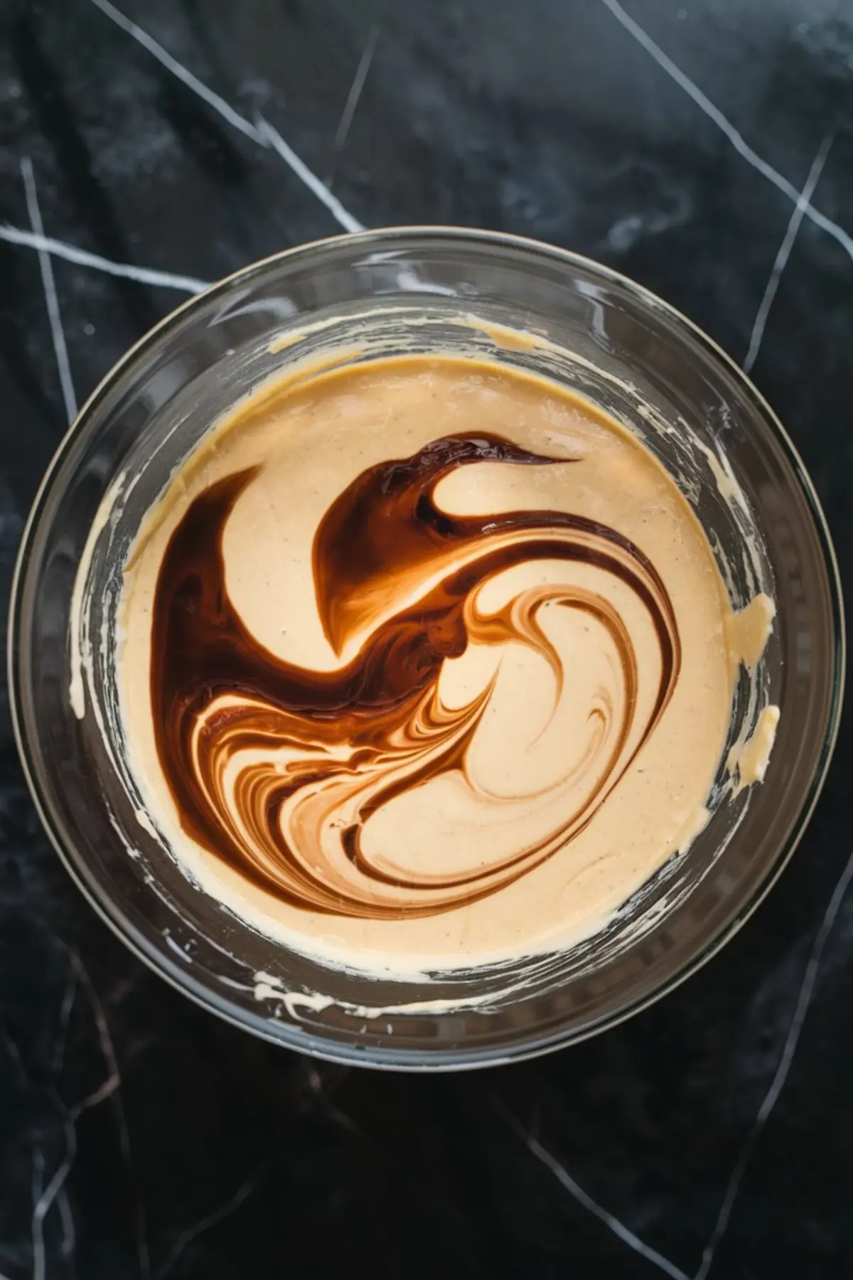 Creamy cheesecake batter swirled with rich espresso mixture in a glass bowl, creating a marbled coffee pattern on a dark marble countertop.
