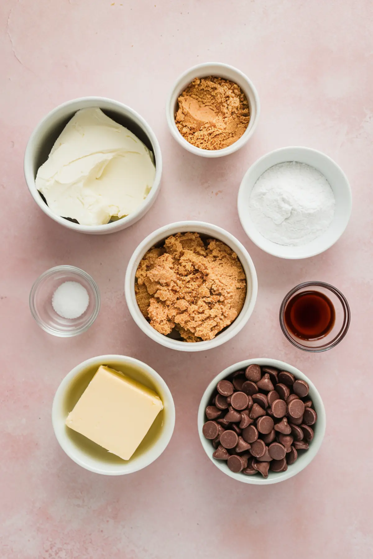Flat lay of small bowls containing cookie dough dip ingredients including cream cheese, brown sugar, powdered sugar, chocolate chips, butter, vanilla, and salt on a pink surface.
