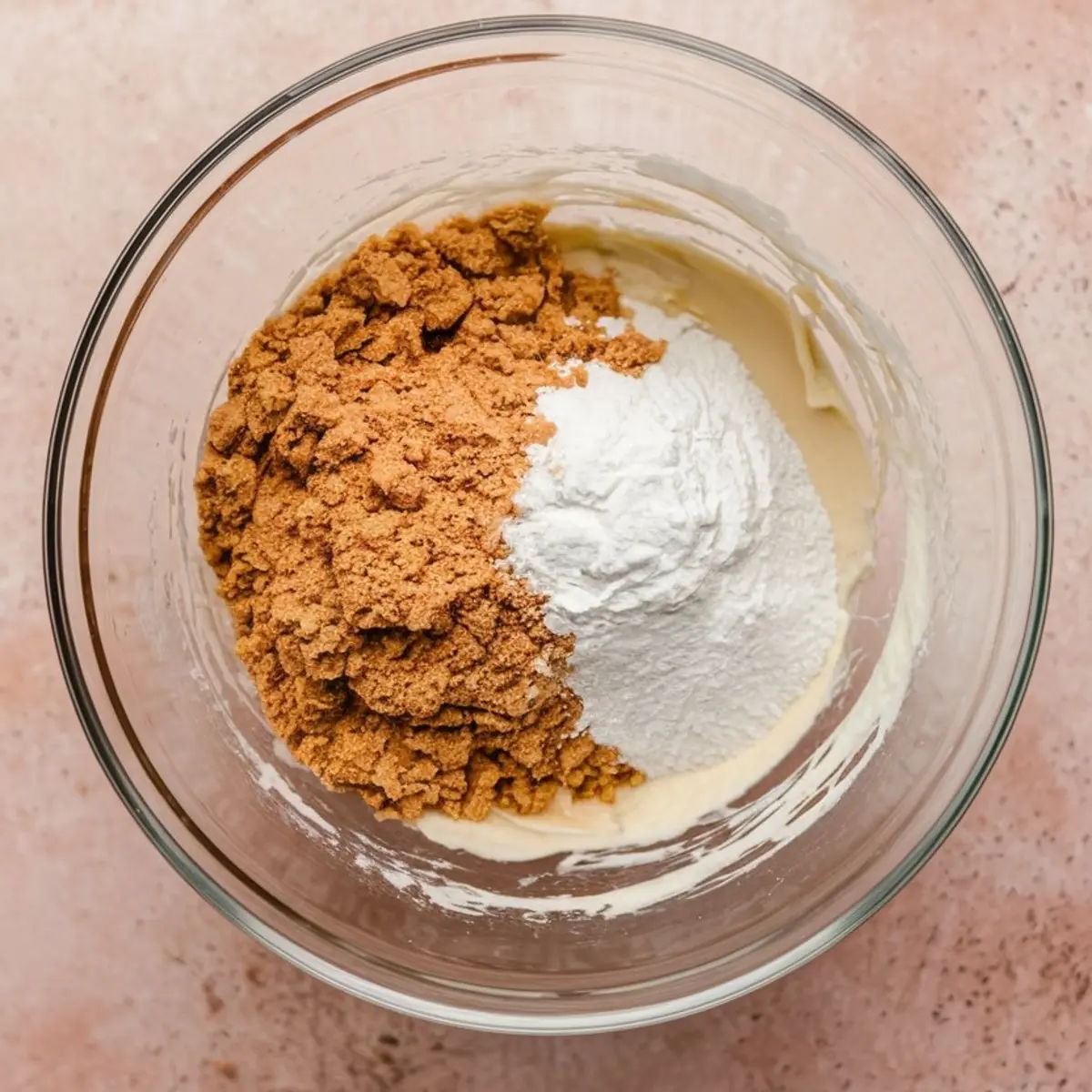 Glass bowl with brown sugar, flour, and white powder ingredients sitting on top of a pale batter, placed on a pink countertop.
