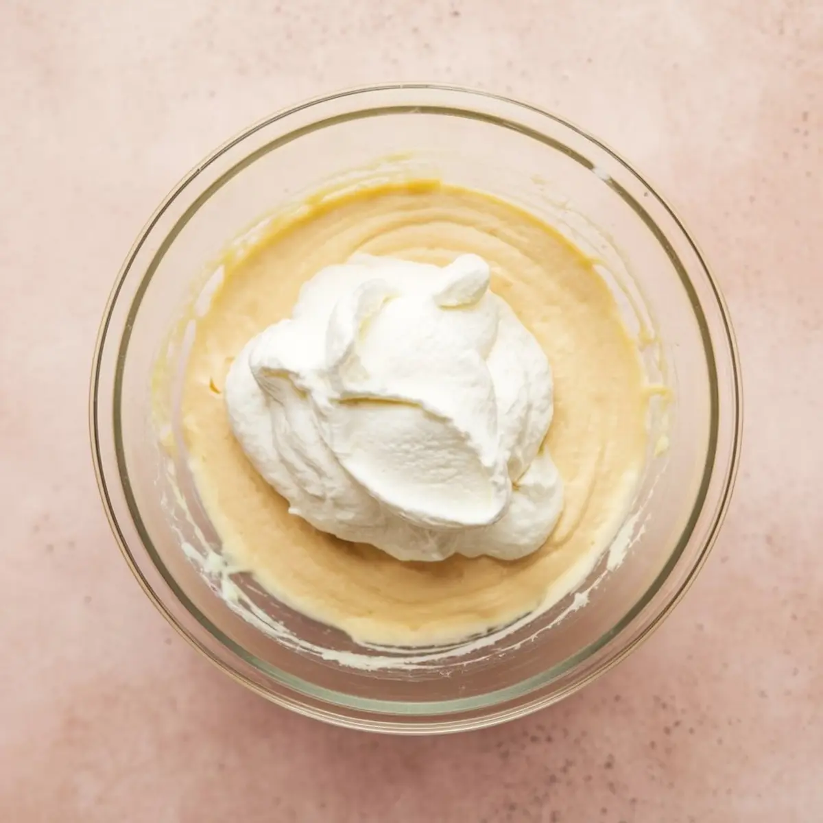 Large dollop of whipped topping added to a bowl of creamy batter, set in a clear glass bowl on a light pink background.
