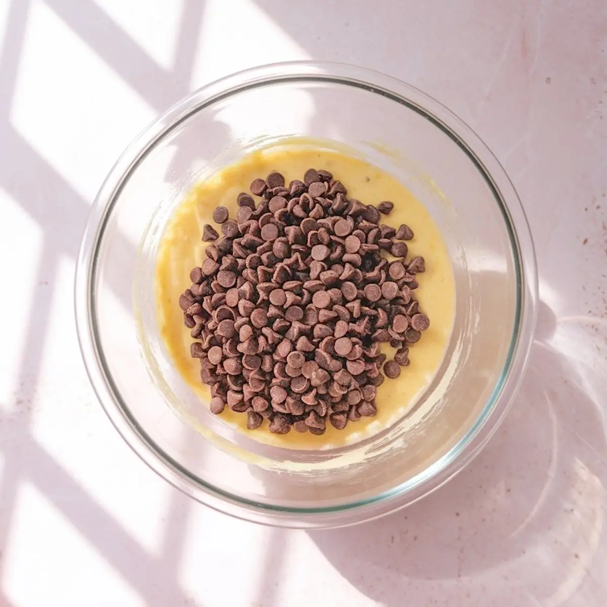 Bowl of cookie dough with a large mound of chocolate chips added on top, placed in natural sunlight with shadows across the surface.
