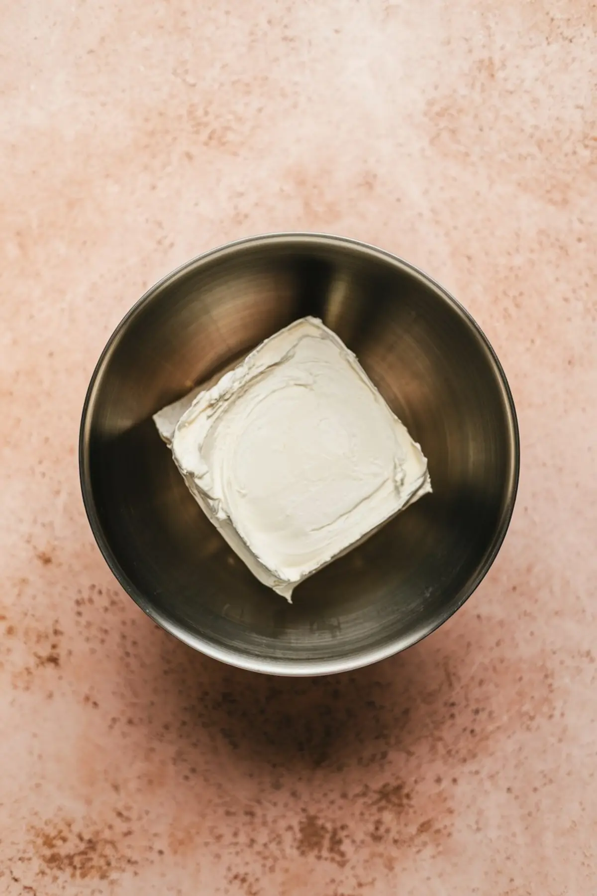 Block of cream cheese in a stainless steel mixing bowl on a pink marbled surface, ready for blending in a dip or frosting recipe.