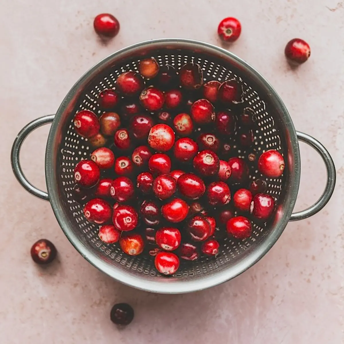 Fresh whole cranberries in a metal colander with handles, set on a light pink surface, showcasing vibrant red fruit ready for cooking.
