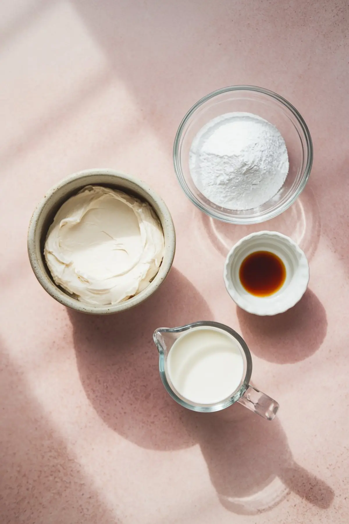 Flat lay of cream cheese dip ingredients including a bowl of cream cheese, powdered sugar in a glass bowl, vanilla extract in a small dish, and heavy cream in a glass measuring cup, all set on a pink background.