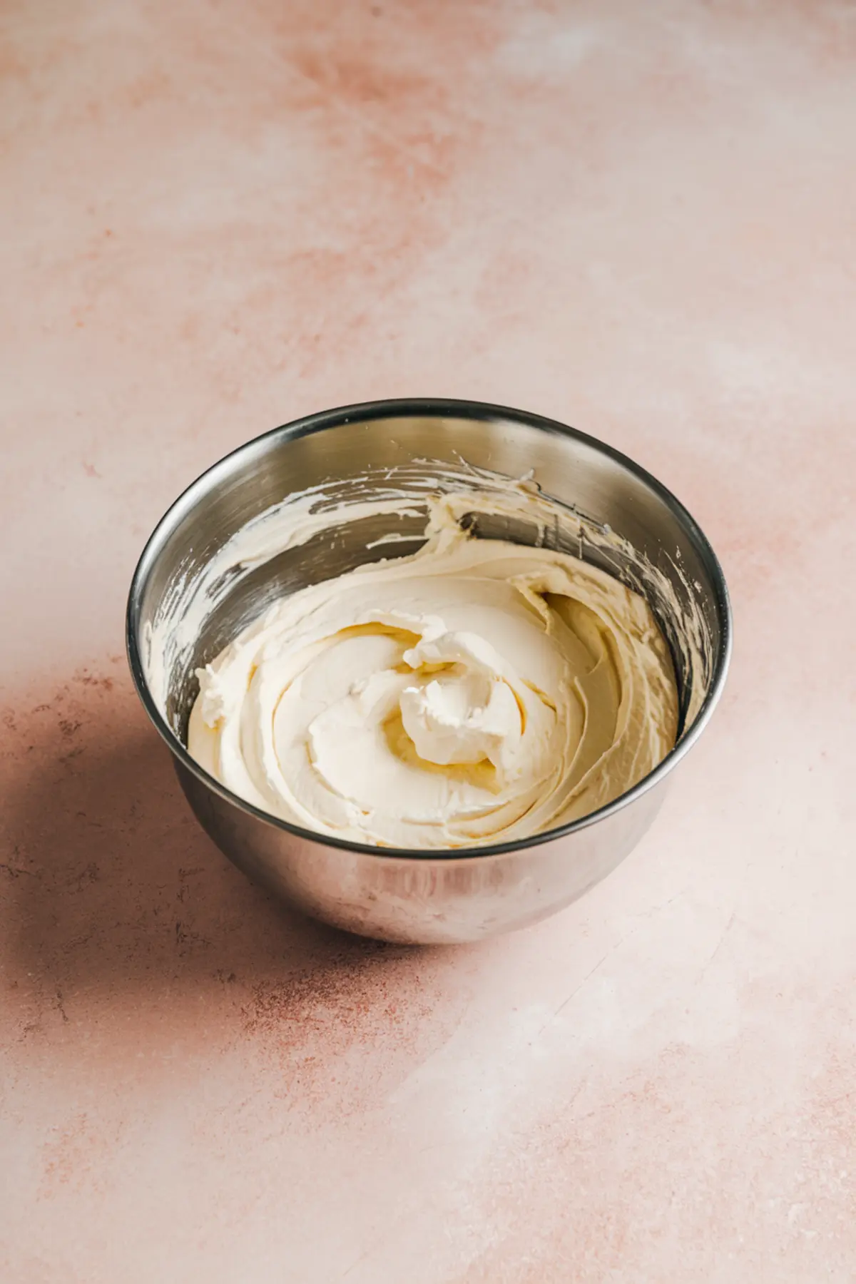 Stainless steel mixing bowl filled with smooth and fluffy cream cheese mixture on a light pink textured background, showcasing creamy texture for dessert dip or frosting recipes.