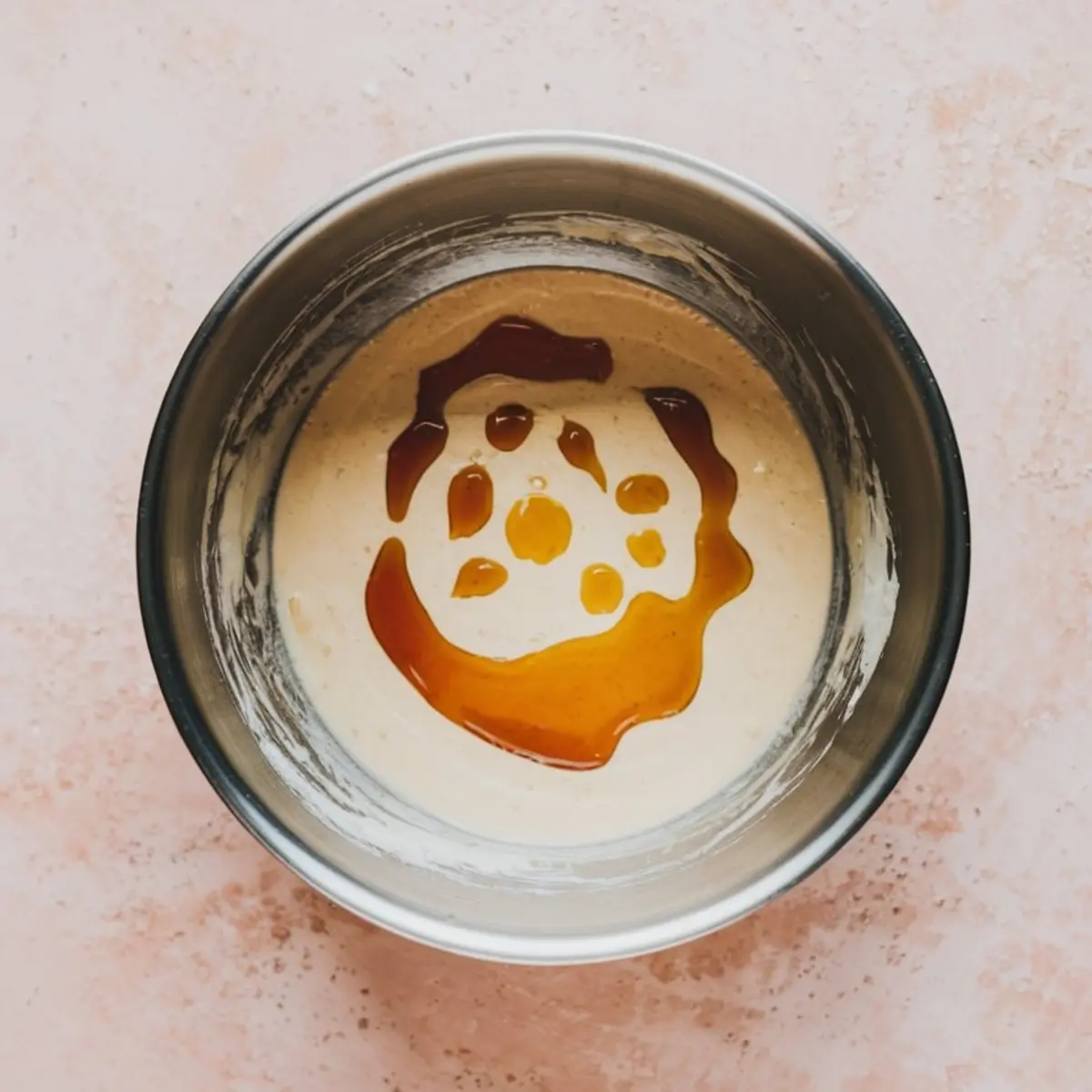 Overhead view of a metal mixing bowl with creamy batter topped with swirls of golden honey and droplets of lemon juice, placed on a pink surface, showing a preparation step for cream cheese dip.