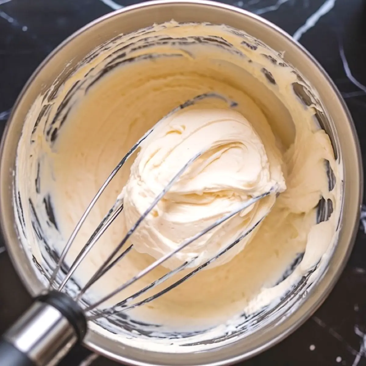 Whipped cream cheese mixture in a stainless steel mixing bowl with a balloon whisk, highlighting the thick, velvety texture essential for making eggnog cheesecake batter.