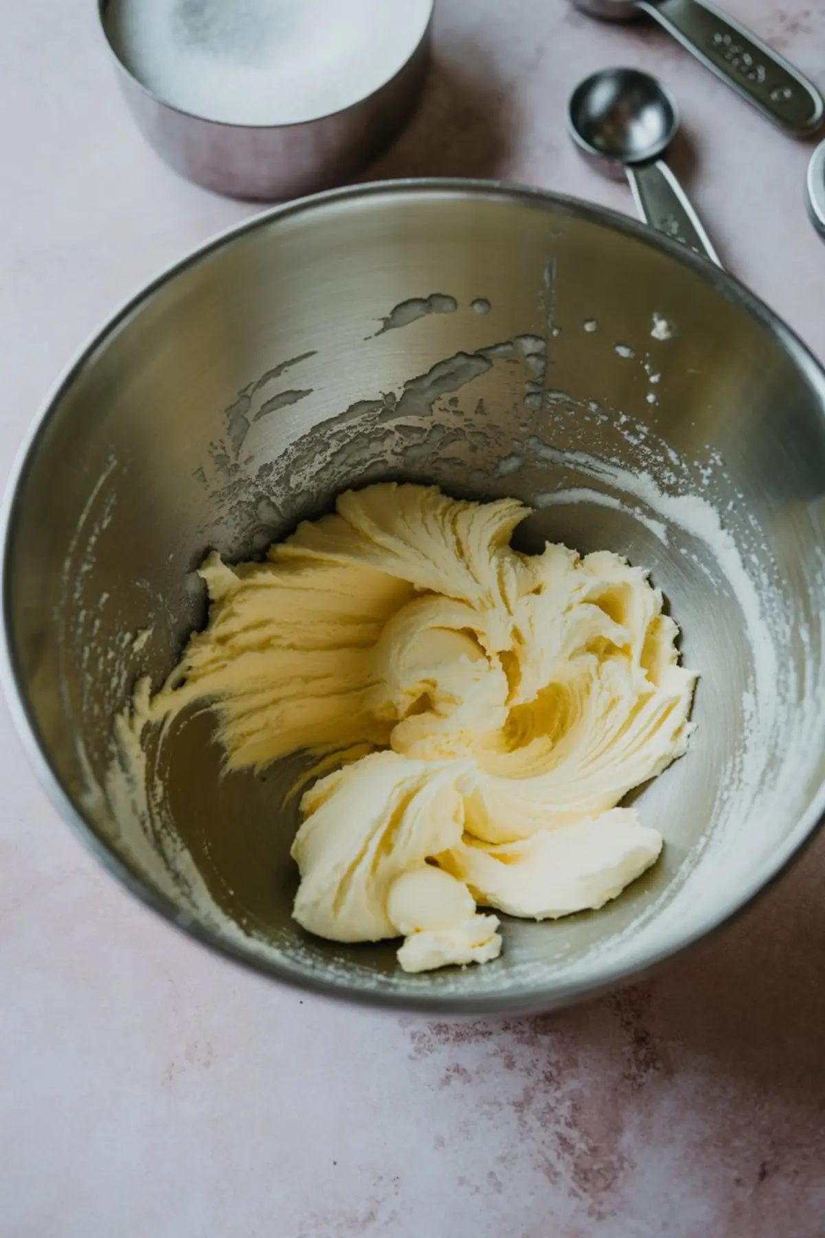 Creamed butter mixture in a metal mixing bowl, partially blended with sugar, shown with measuring cups and spoons on a light pink surface.