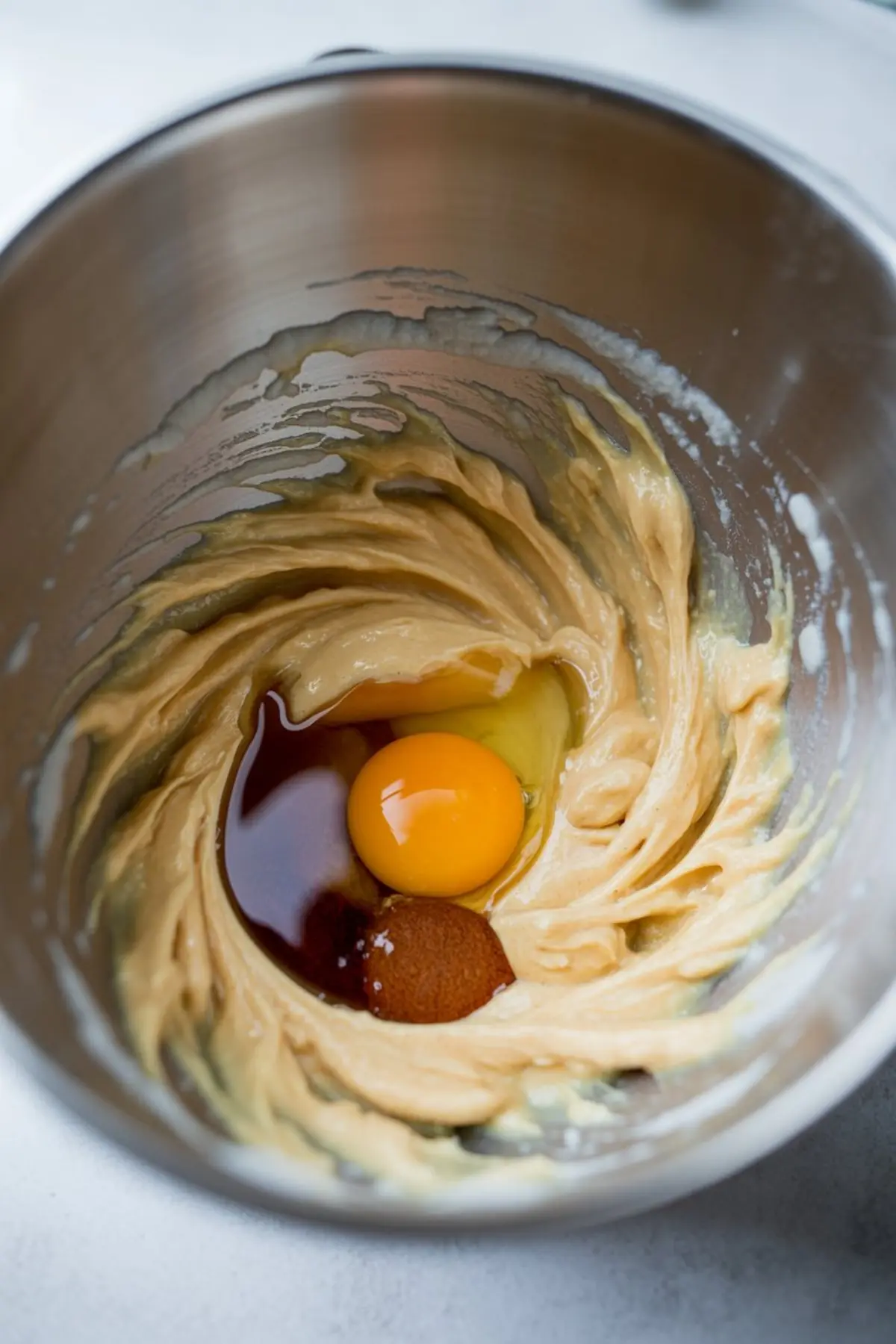 Mixing bowl containing creamed butter with added egg, brown sugar, and vanilla extract, ready for blending on a baking prep surface.