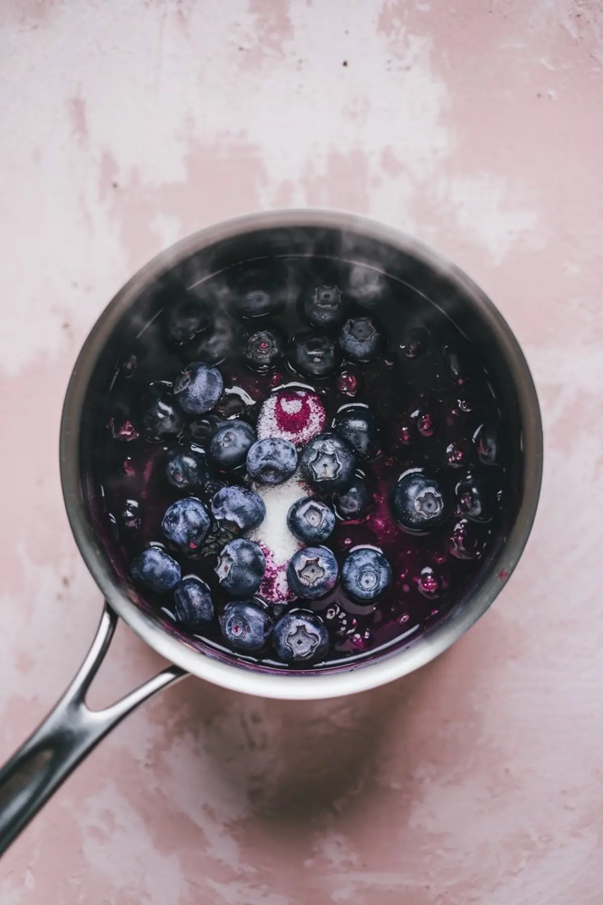 Saucepan with fresh blueberries, sugar, and juices beginning to simmer, showing the start of a blueberry compote on a pink background.