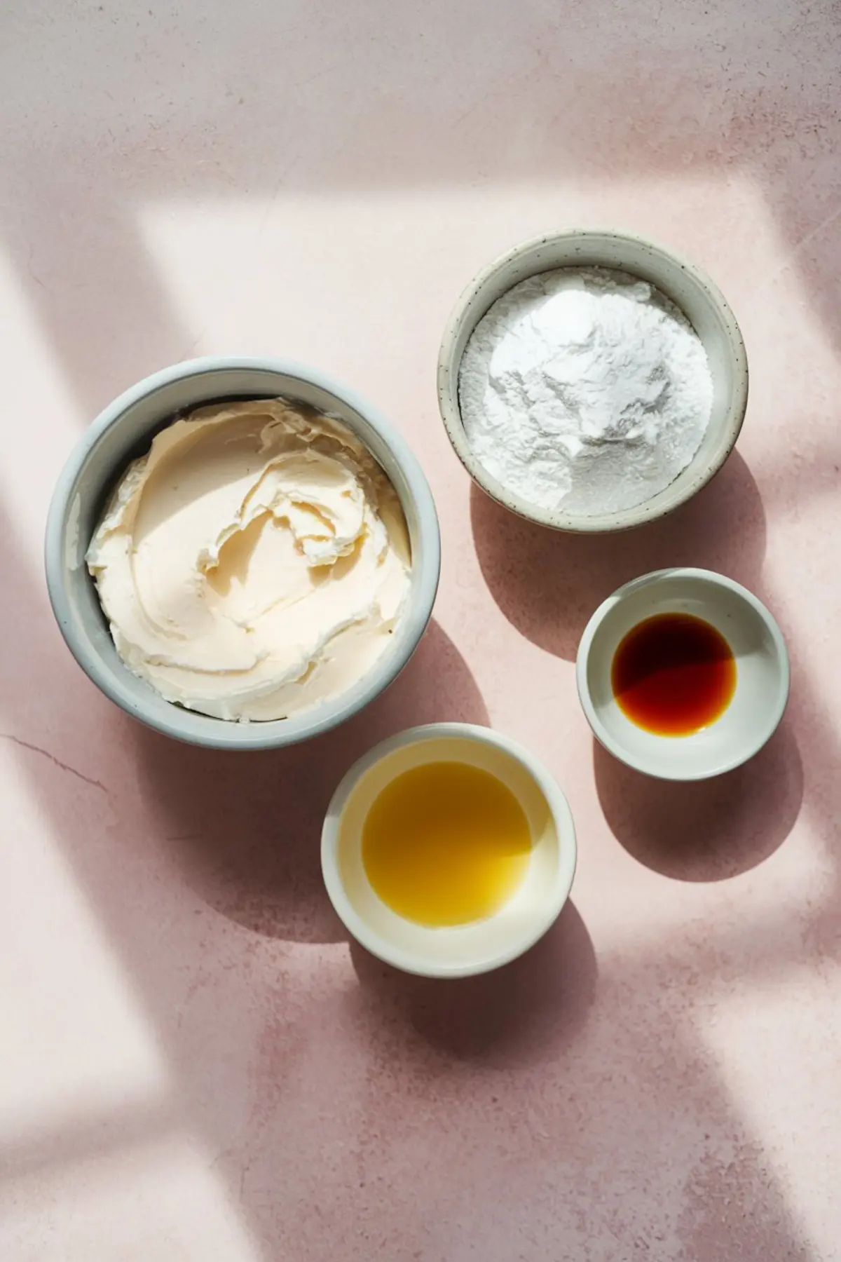 Four small bowls on a pink surface containing cream cheese, powdered sugar, vanilla extract, and orange juice, arranged in soft natural light.

