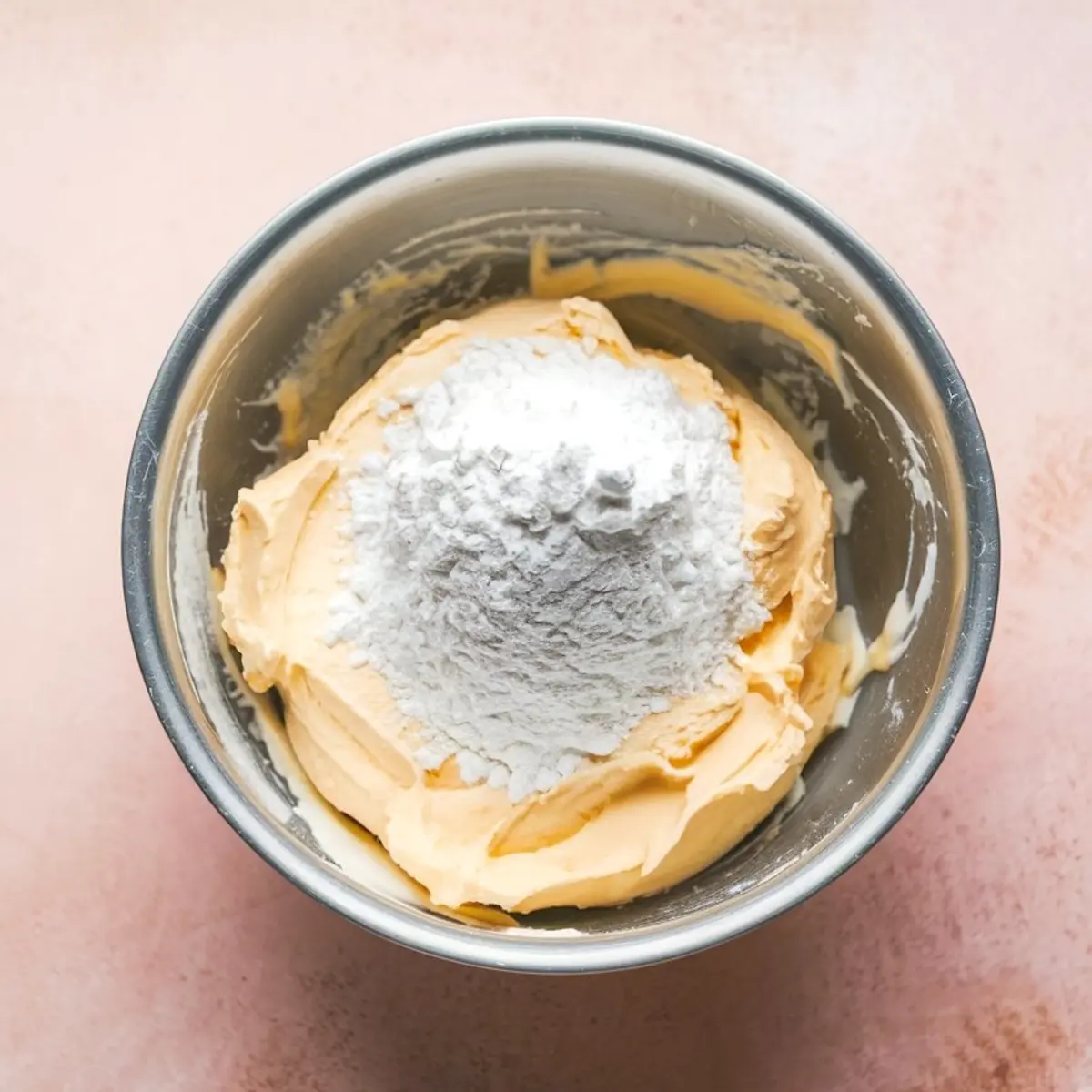 Metal mixing bowl with whipped cream cheese and a mound of powdered sugar on top, set against a light pink background.