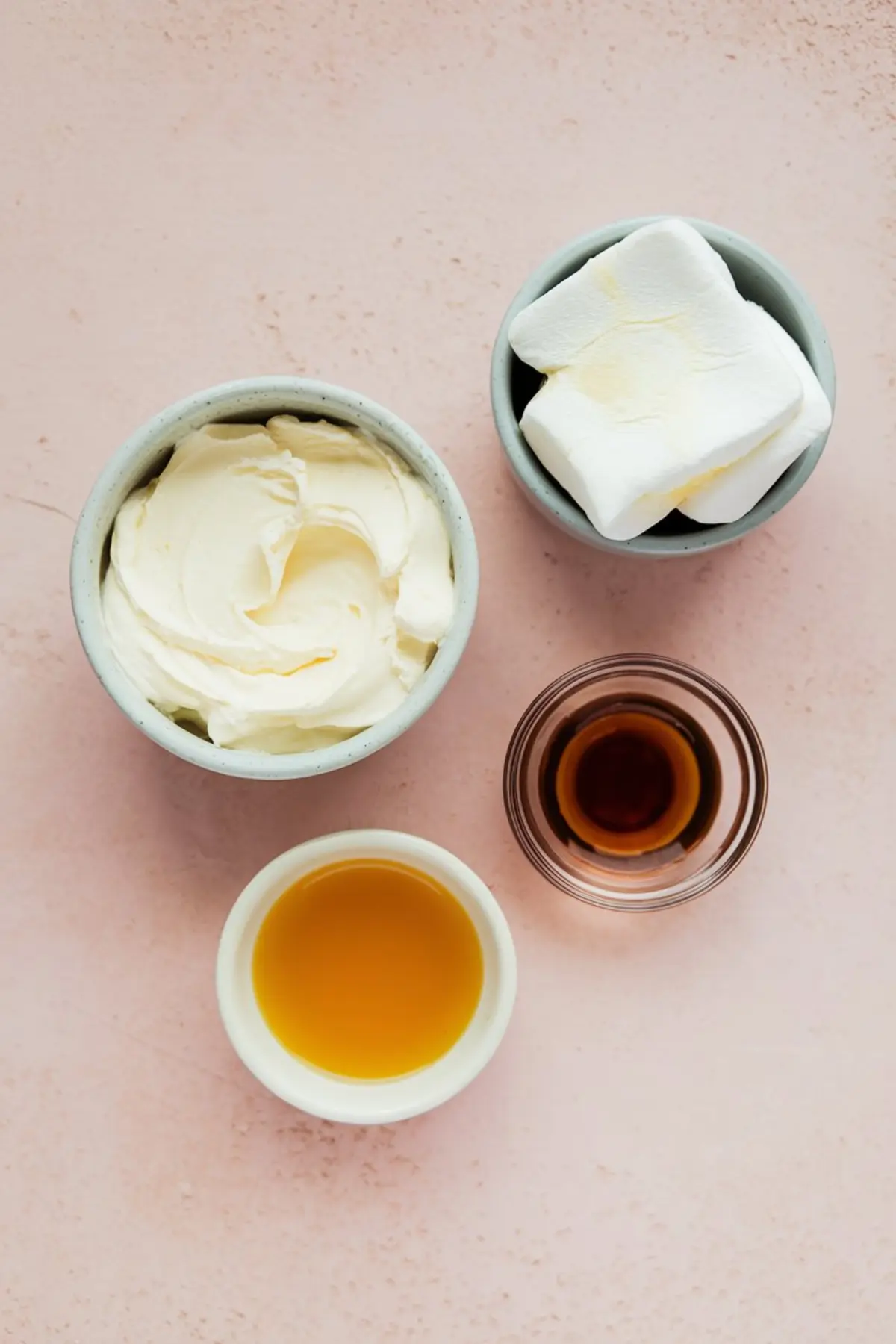 Four small bowls with ingredients for marshmallow fluff fruit dip on a light pink surface. One bowl holds whipped cream cheese, one contains large marshmallows, another has golden honey, and the last contains dark vanilla extract.
