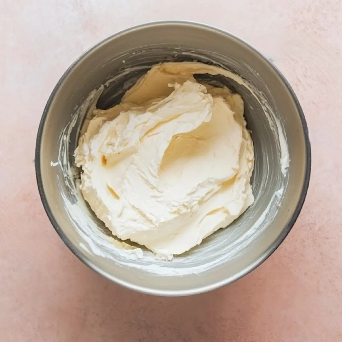 A metal mixing bowl filled with smooth, whipped cream cheese mixture placed on a light pink background, showing the creamy texture of the dip base.
