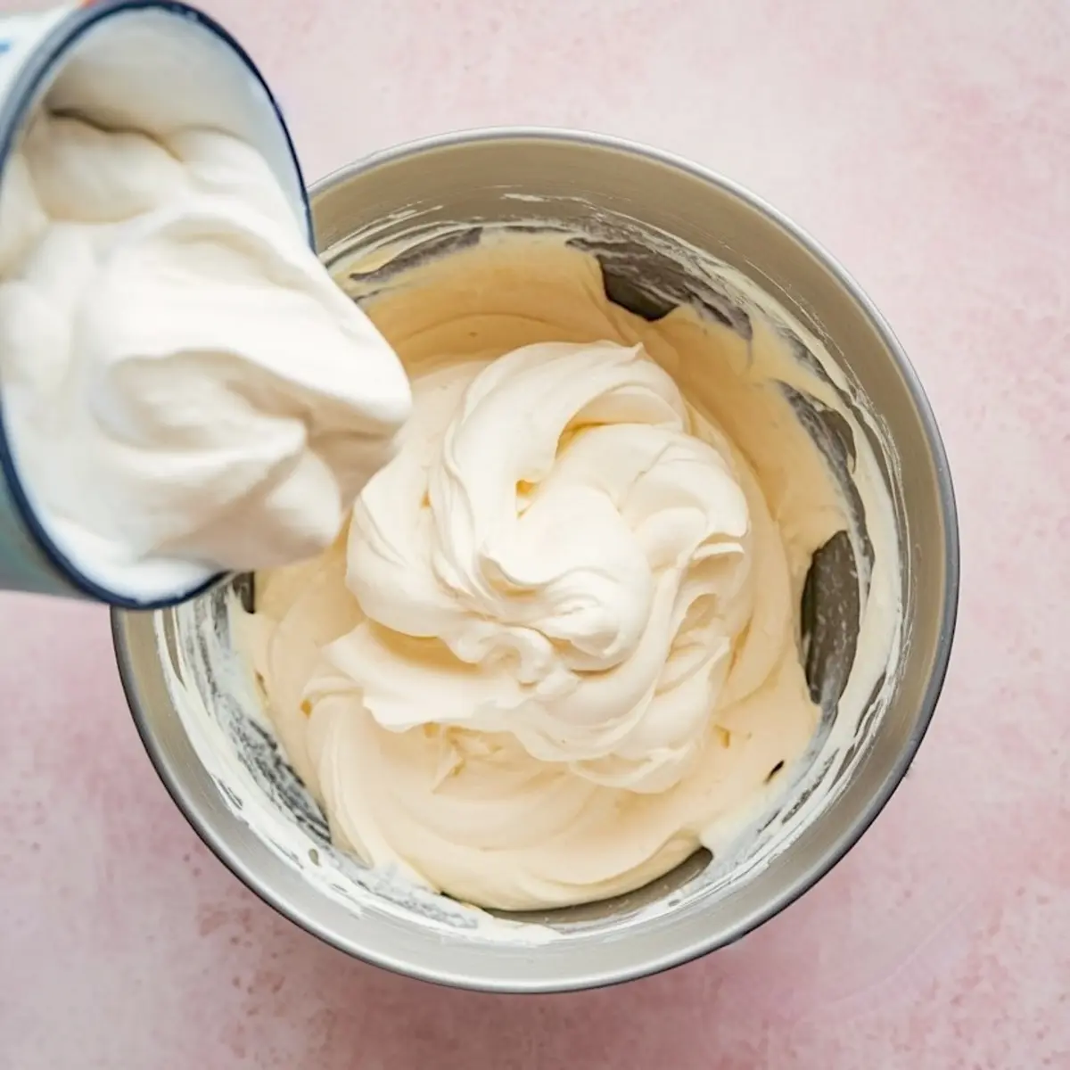 Whipped mixture being poured from a container into a mixing bowl filled with fluffy cream, demonstrating the preparation of marshmallow fluff fruit dip.
