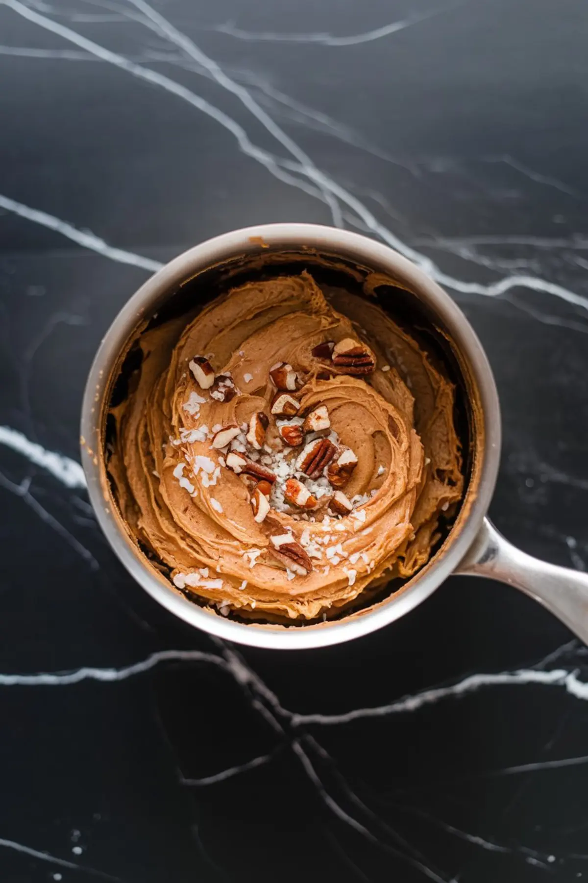Saucepan filled with creamy German chocolate frosting, topped with chopped pecans and shredded coconut, set on a black marble background.