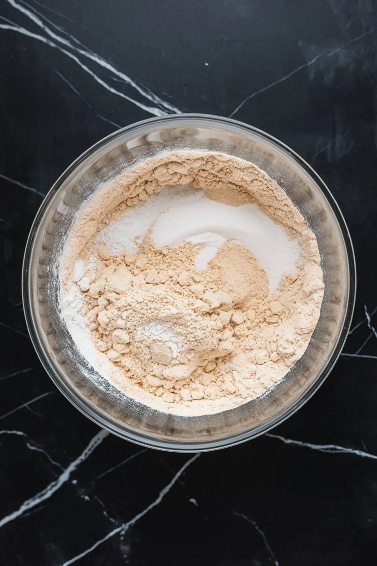 Close-up of a glass mixing bowl filled with blended dry ingredients for baking, featuring flour, sugar, and leavening agents on a black marble surface.