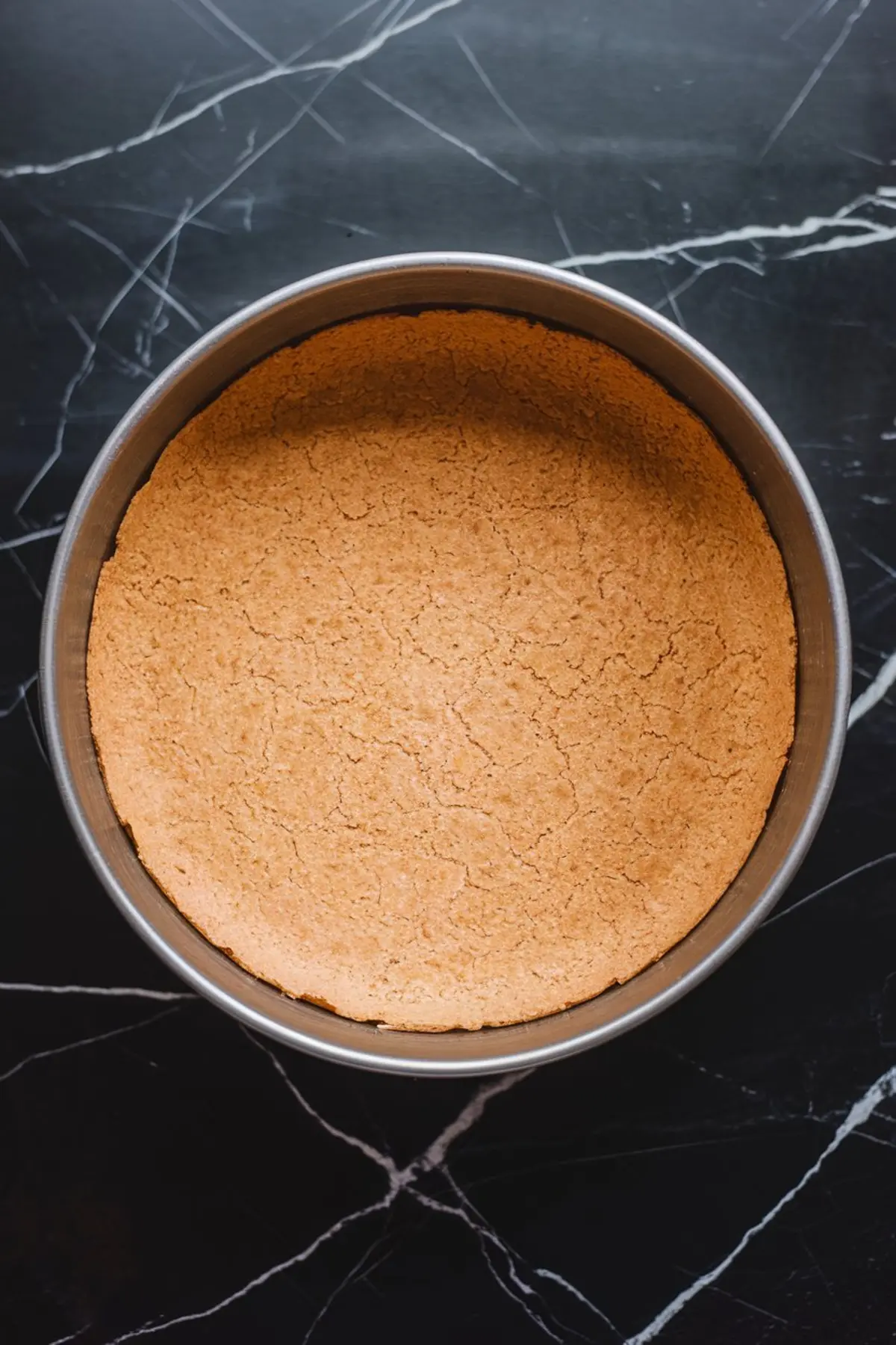 Golden brown baked graham cracker crust in a round springform pan, featuring a firm, even surface on a black marble background, prepared for cheesecake filling.