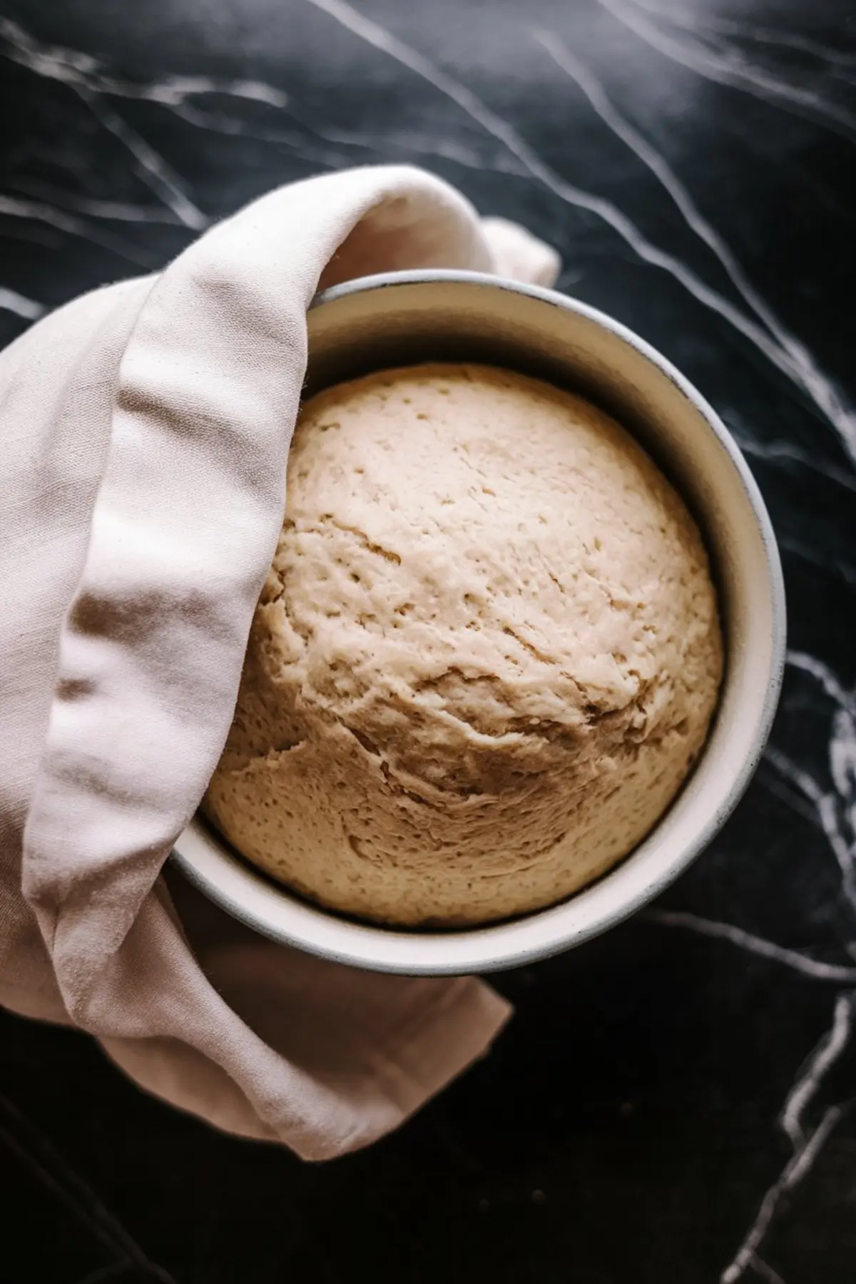 Risen dough in a metal mixing bowl, partially covered with a light cloth, resting on a dark marble countertop.