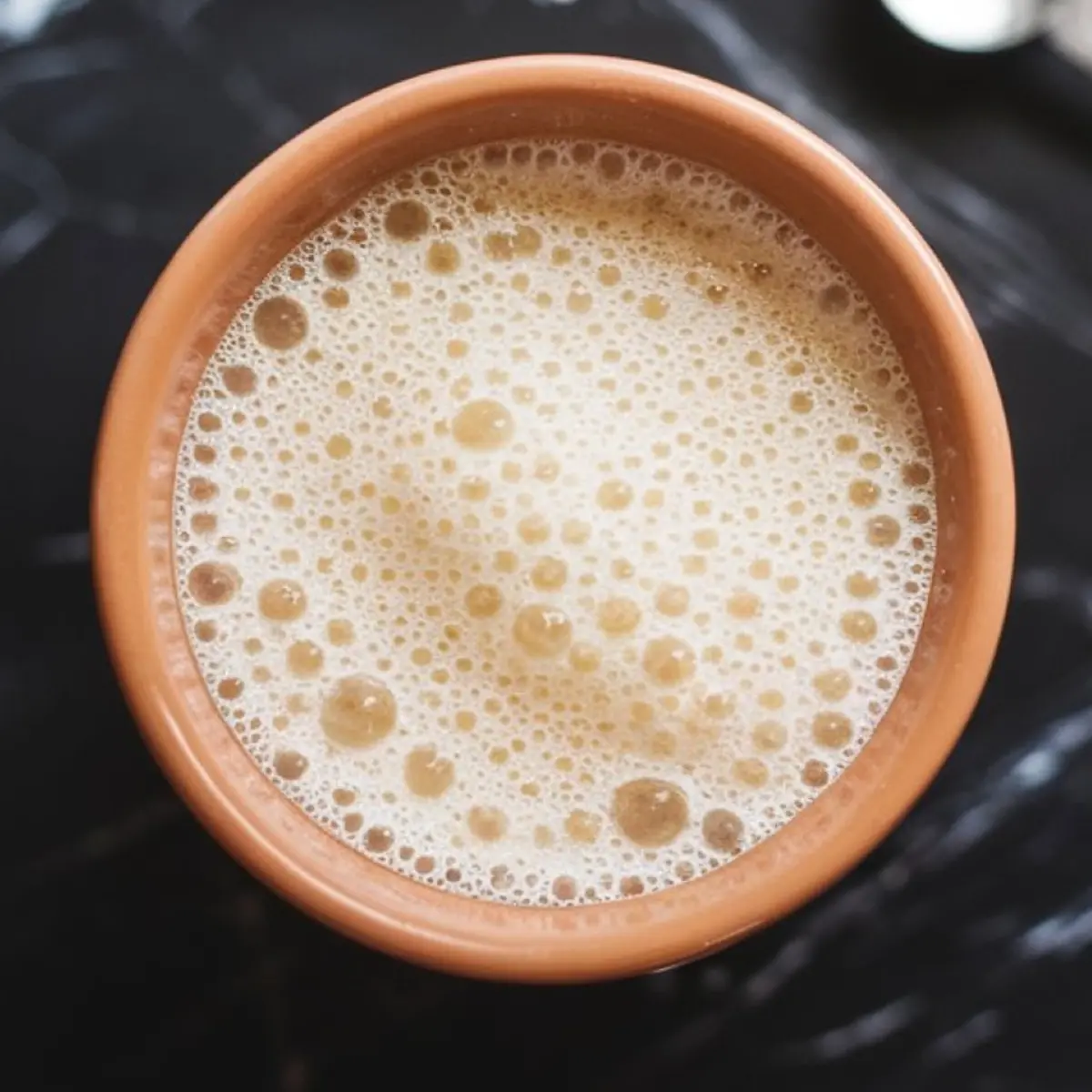 Frothy yeast mixture in a terracotta cup, showing activated bubbles on the surface, placed on a black marble surface.
