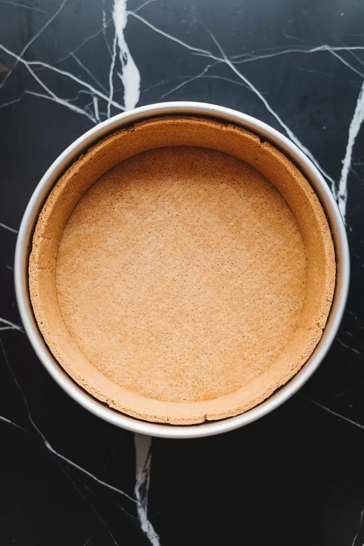 A round graham cracker crust pressed into a springform pan, evenly shaped with smooth sides and bottom, ready for cheesecake filling on a black marble countertop.
