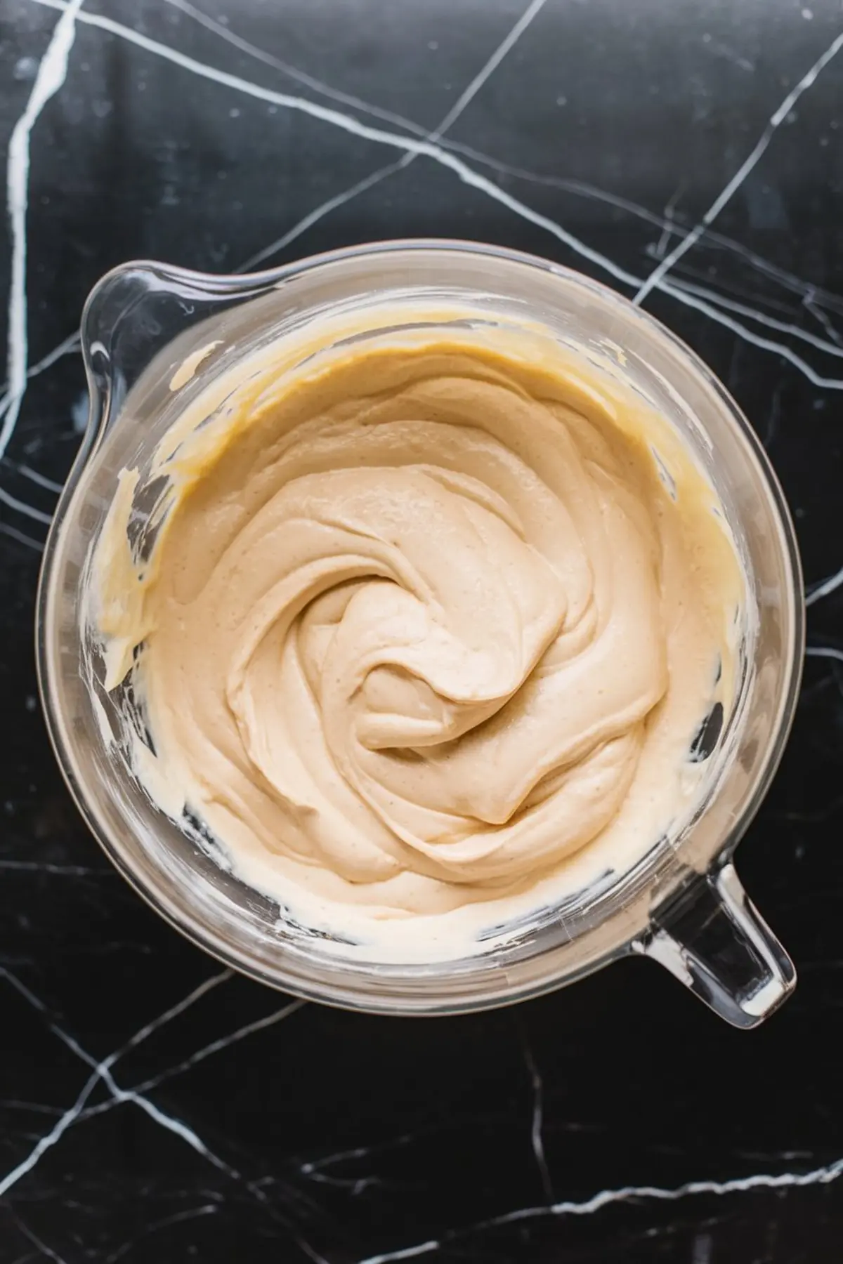 A clear glass mixing bowl filled with thick, creamy cheesecake batter, swirled smoothly and ready for pouring, sitting on a dark marble countertop.
