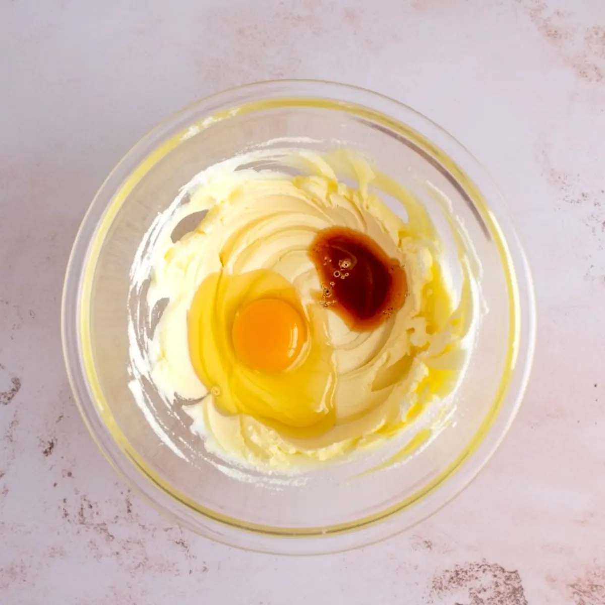 Clear glass bowl with creamed butter and sugar, topped with a raw egg and a splash of vanilla extract, set on a light pink background for a cookie dough base.