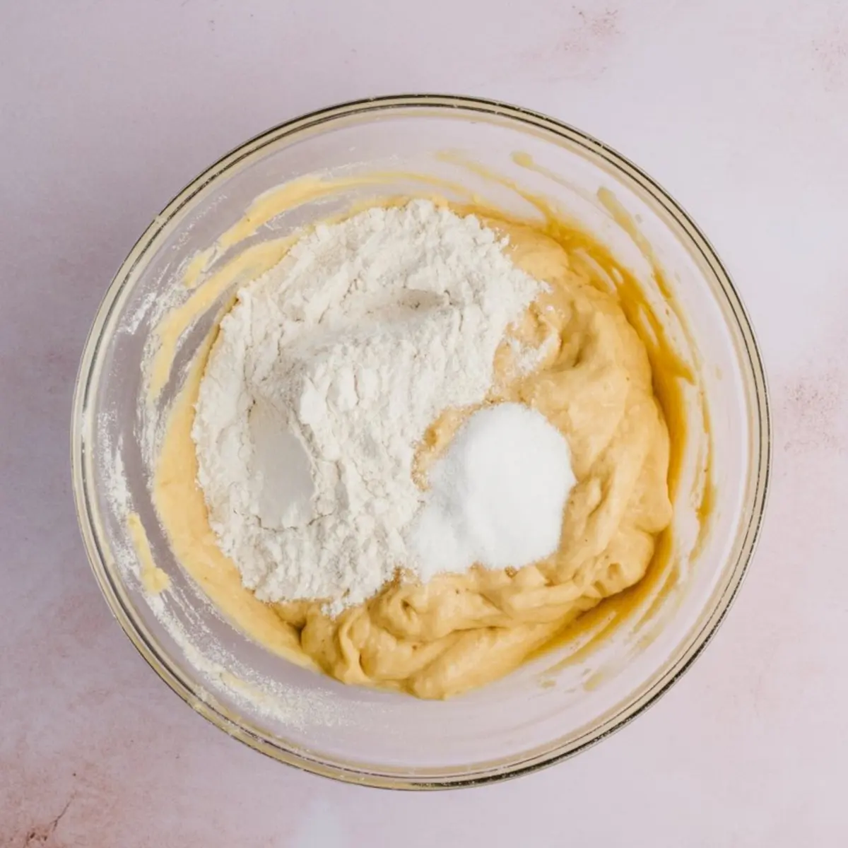 Glass mixing bowl with thick yellow batter, topped with a mound of white flour and a scoop of sugar, ready to be blended for baking.