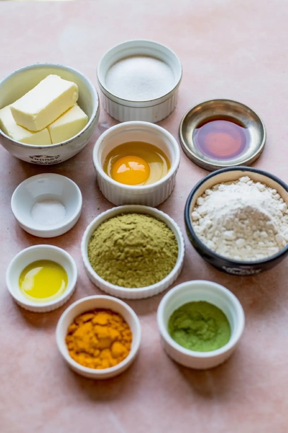 Flat lay of baking ingredients including butter, sugar, egg, flour, matcha powder, mango powder, vanilla extract, and oil, arranged in small bowls on a pink surface.