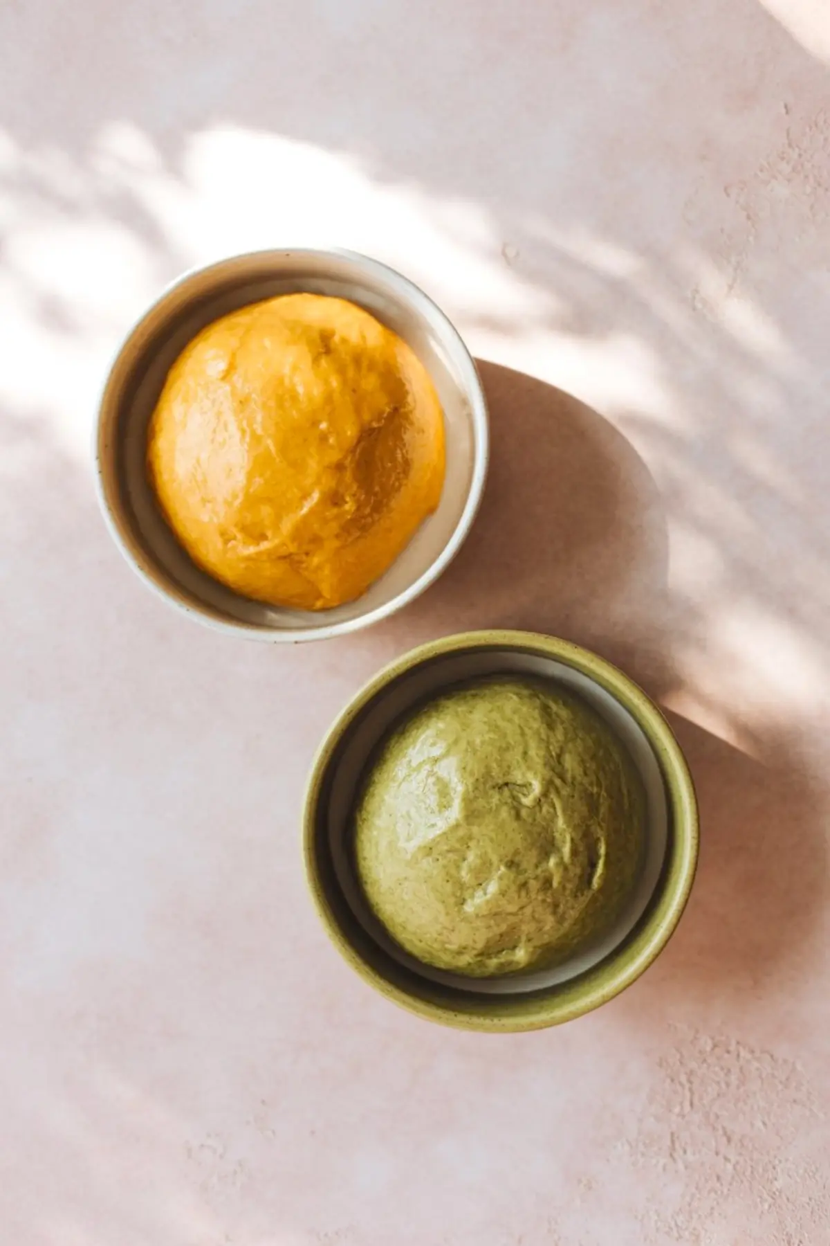 Two ceramic bowls with smooth, colored cookie dough—one bright orange and the other earthy green—representing mango and matcha flavors.