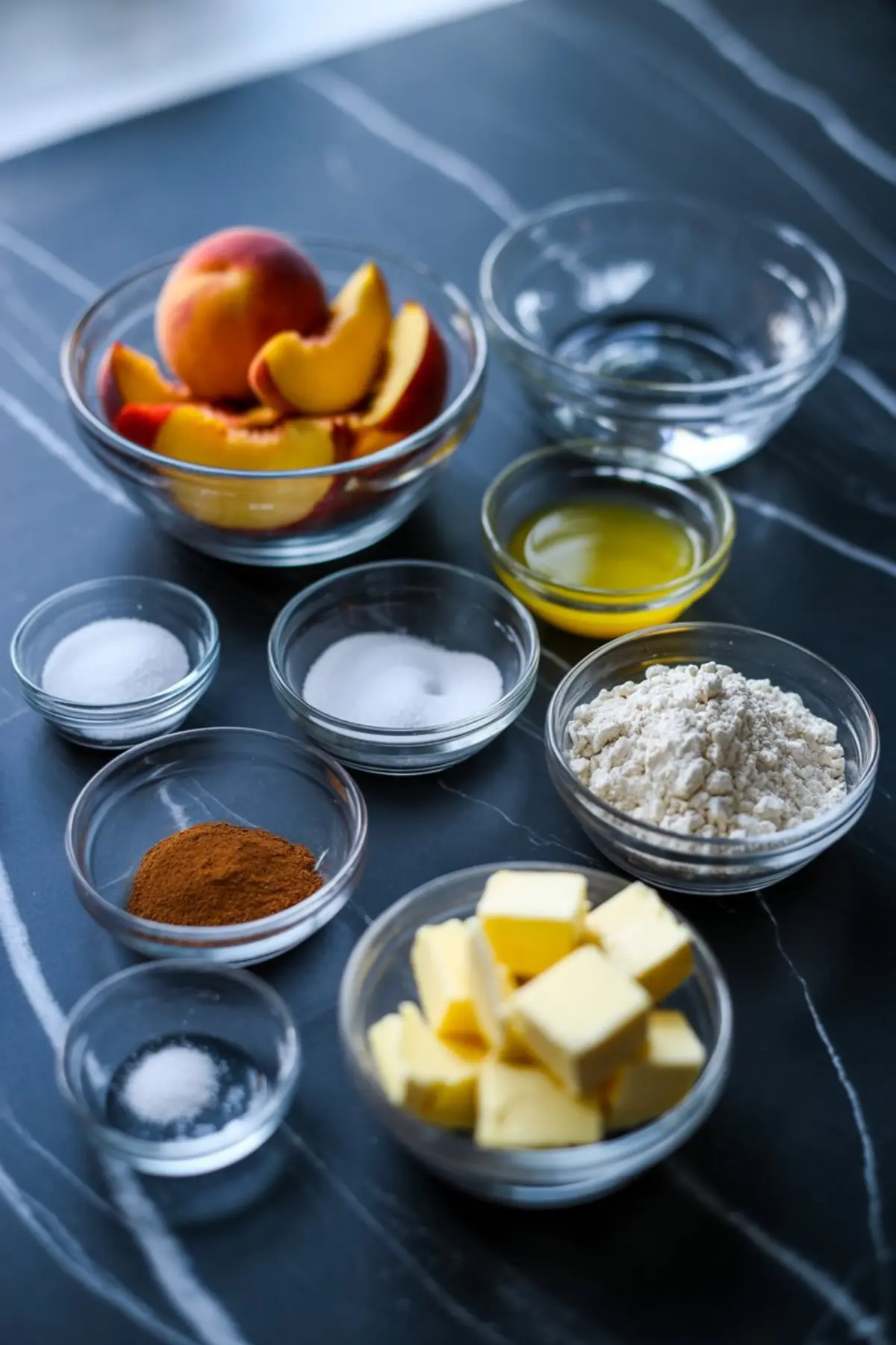 Overhead view of small glass bowls filled with peach pie ingredients including sliced fresh peaches, cubed butter, flour, cinnamon, sugar, salt, and melted butter on a dark marble countertop.