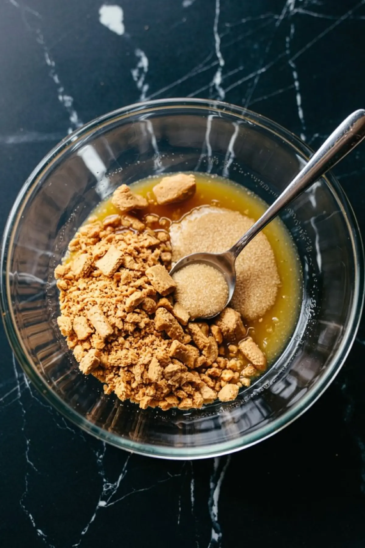 Clear glass mixing bowl with crushed graham crackers, granulated sugar, and melted butter, prepared for making cheesecake crust, placed on a black marble countertop.
