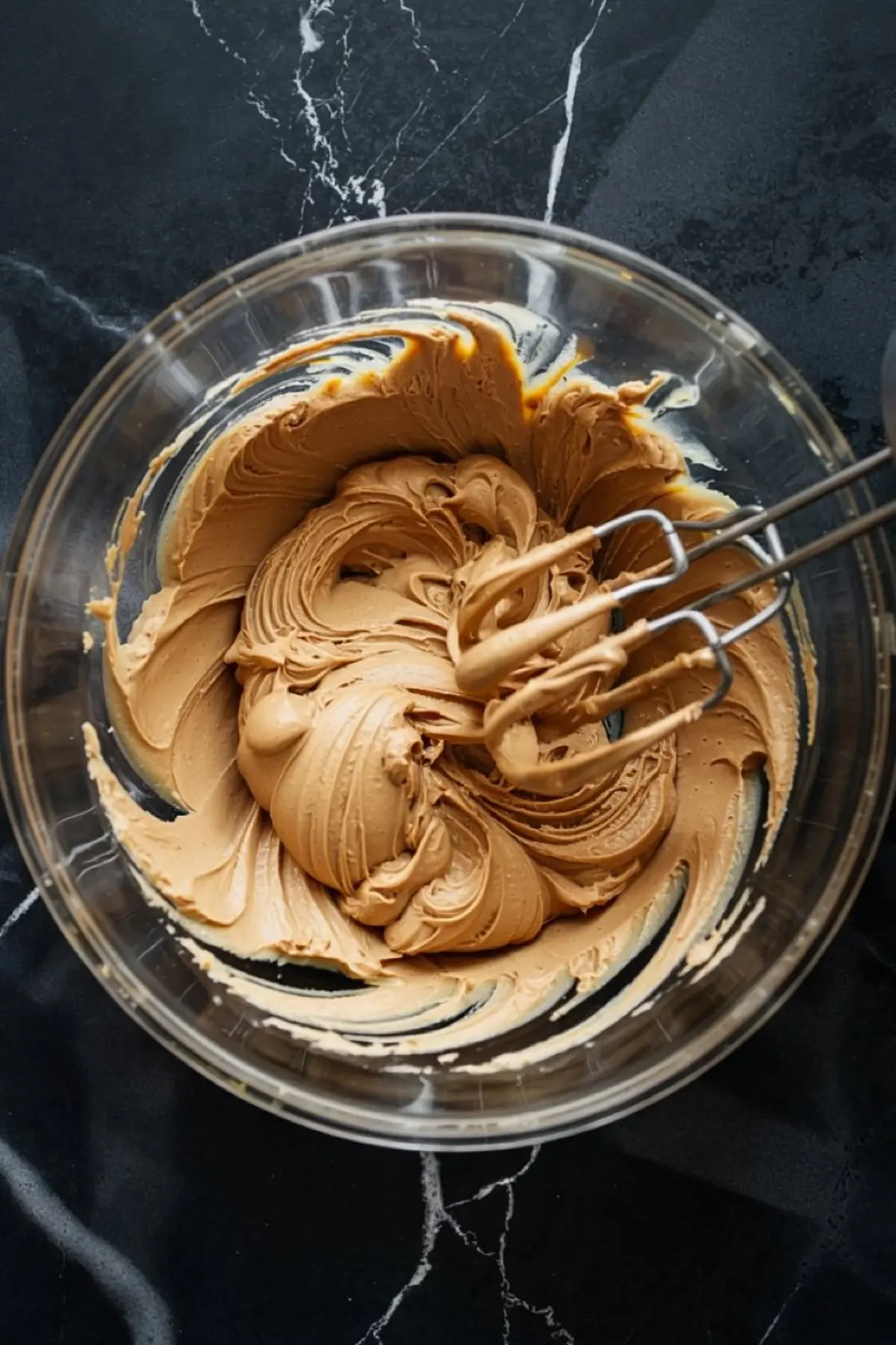 Glass bowl filled with creamy peanut butter mixture being whipped with metal beaters, creating smooth and fluffy cheesecake filling on a black marble surface.
