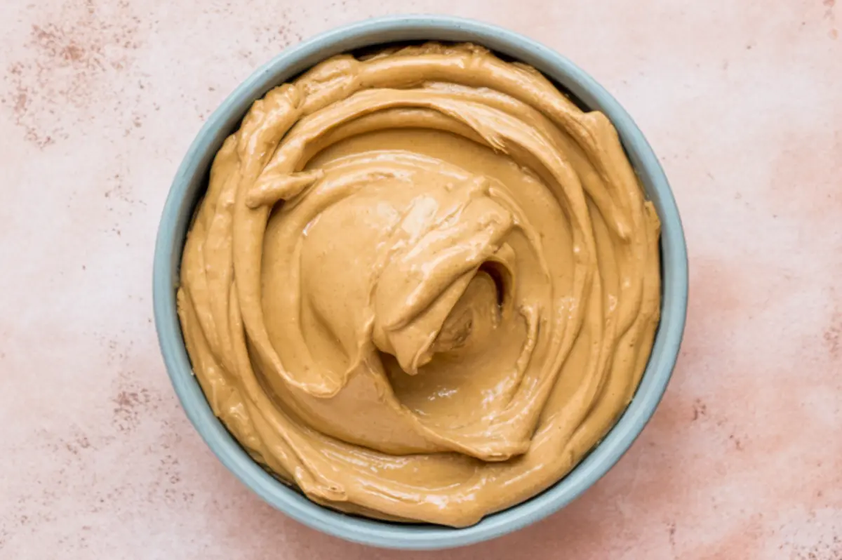 Velvety peanut butter dip in a blue bowl with a glossy, creamy swirl pattern on a pale pink background.