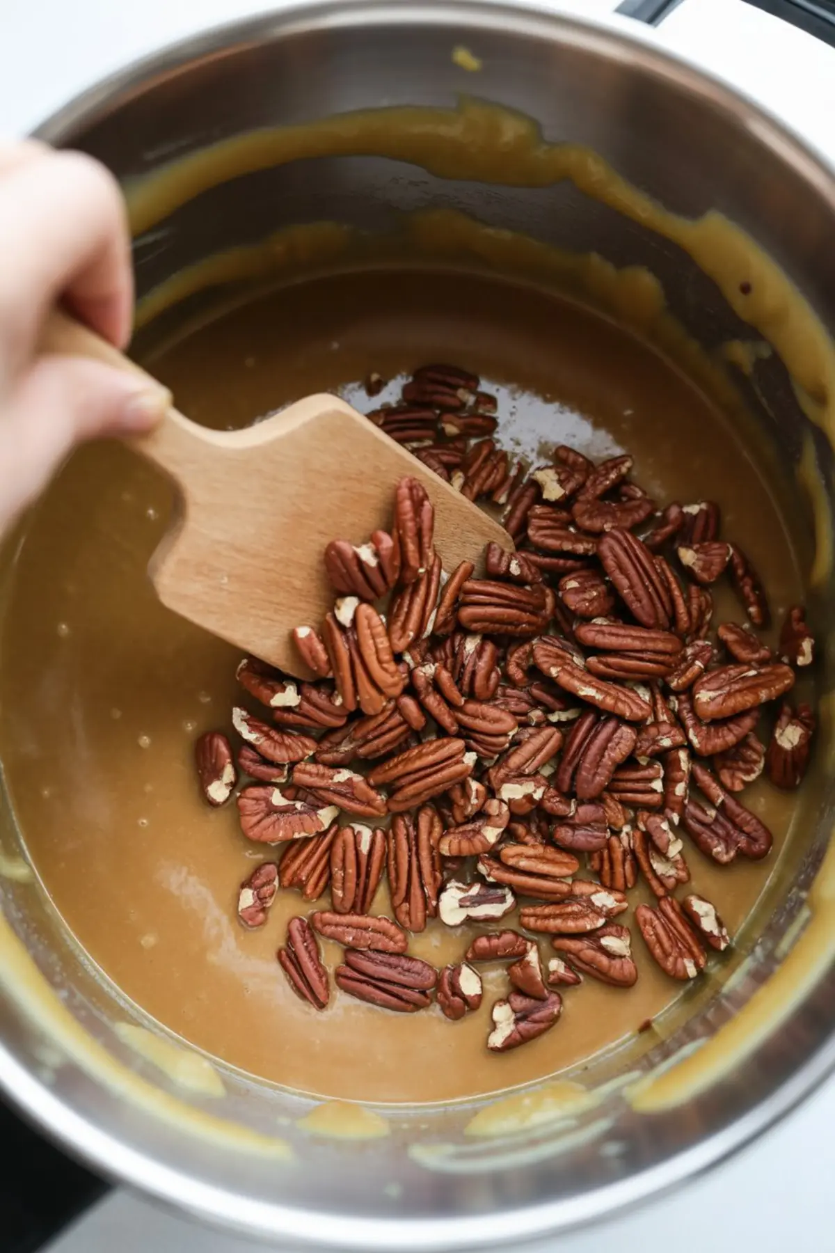Whole pecans being stirred into a thick caramel-like brown sugar and butter mixture in a metal pot, showing an early step in making pecan pie dip on the stove.
