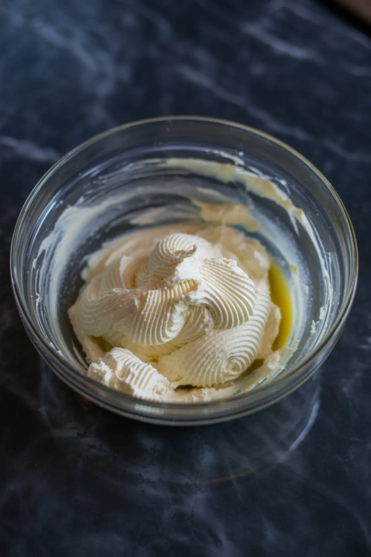Piped swirl of cream cheese filling in a glass bowl on a dark marble counter, showing thick, whipped texture ready for muffin filling.
