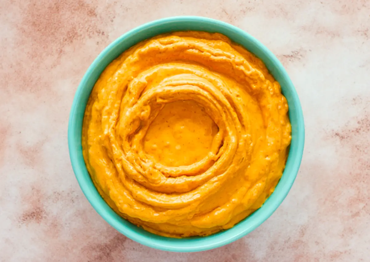 Overhead view of a teal bowl filled with smooth, whipped pumpkin pie dip, swirled with a spoon on a light marbled background.