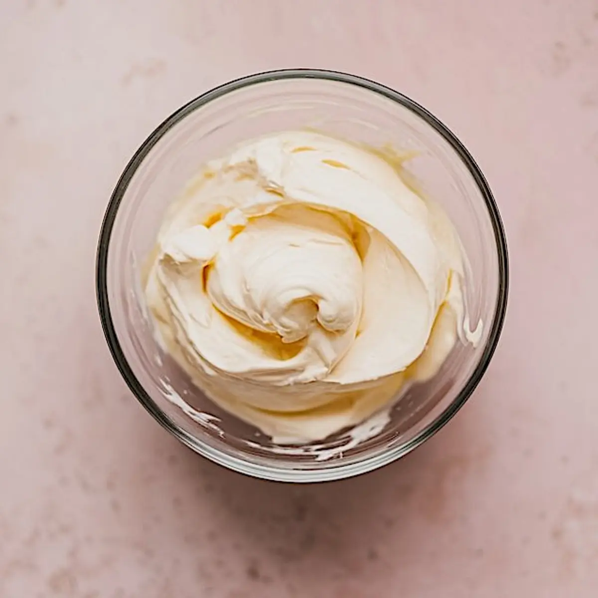 Smoothly whipped cream cheese mixture in a glass bowl, prepared on a pink countertop.